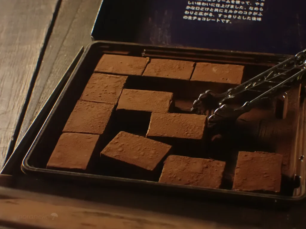 木目のテーブルの上で、開封されたロイズ生チョコレート［オーレ］のトレイに整然と並ぶ、ココアパウダーをまとった四角い生チョコ。右側からシルバーのトングで一粒を持ち上げようとしている。