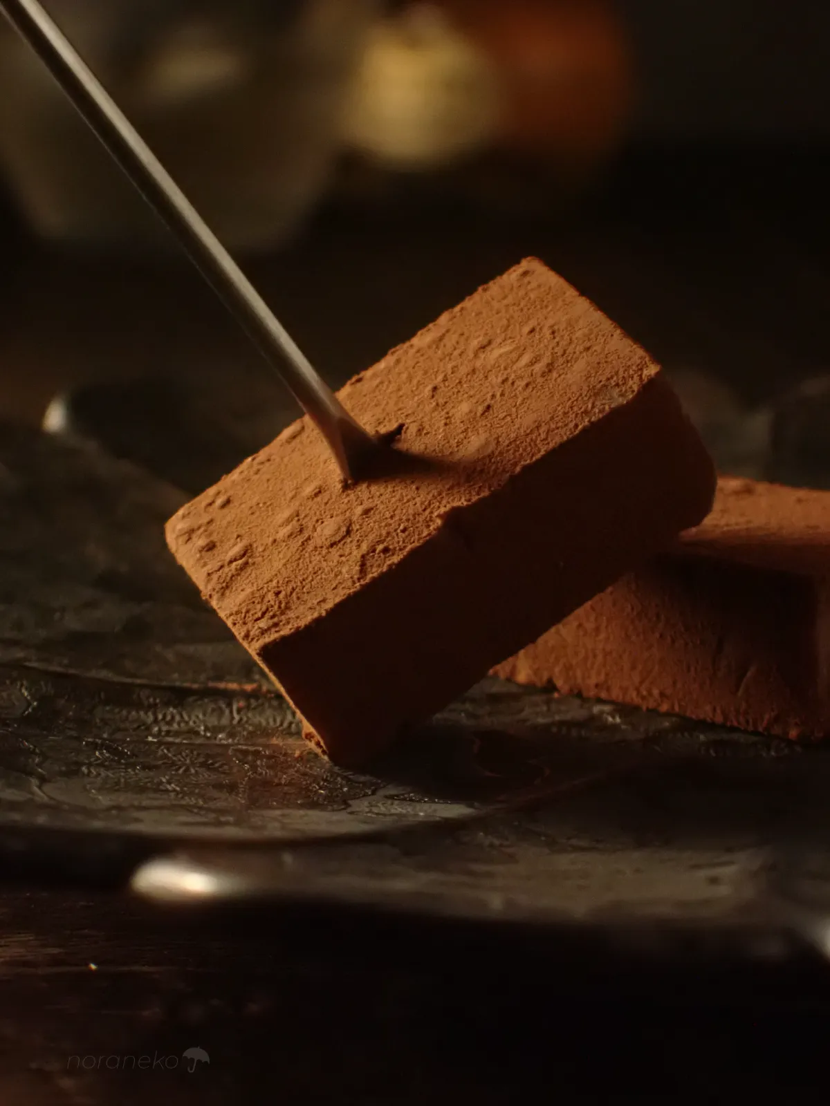 器に載ったロイズ生チョコレート［オーレ］の一粒に、シルバーのピック（トング）が刺さっている。柔らかいチョコにピックが沈み込む様子が捉えられている。