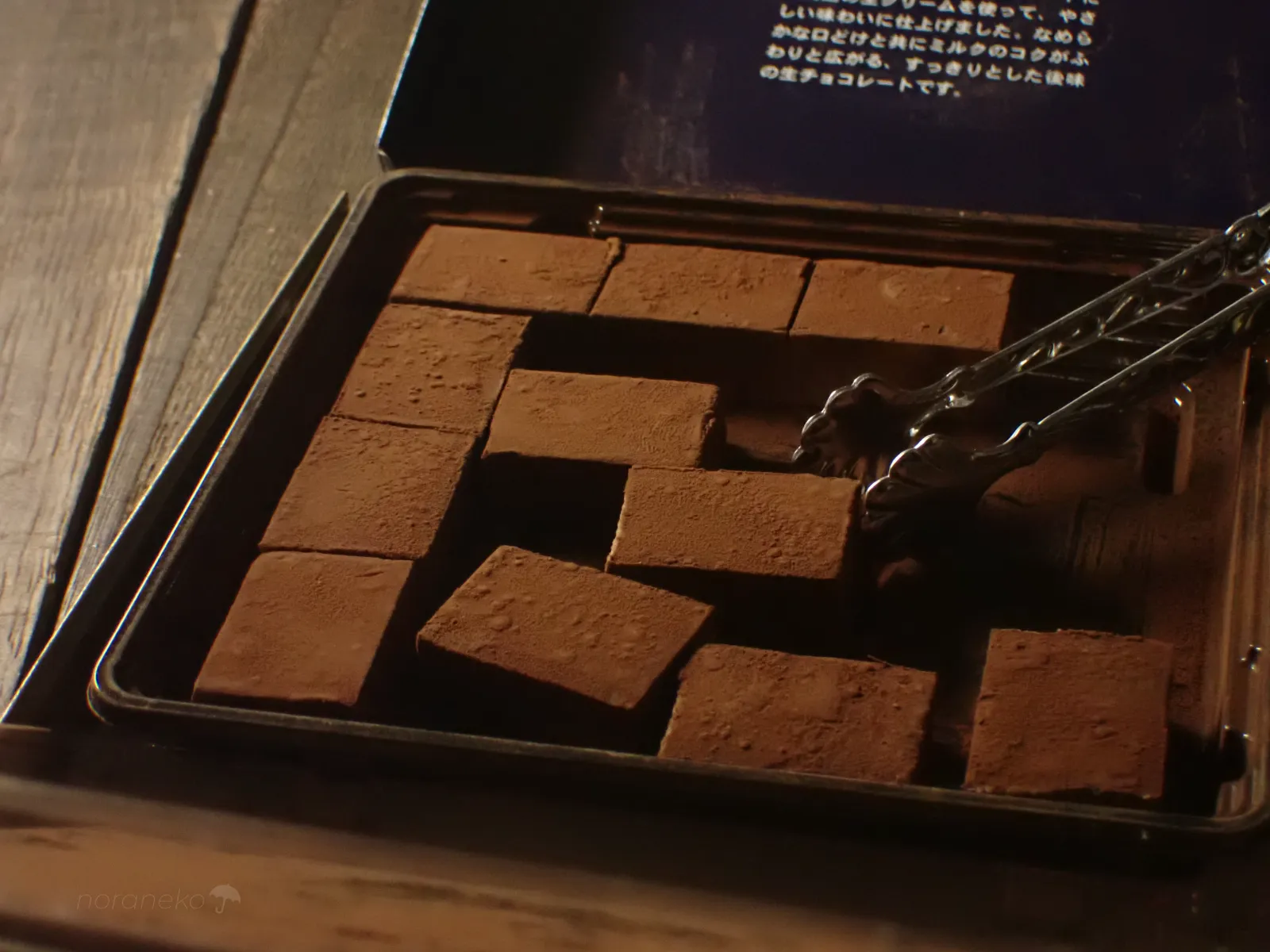 木目のテーブルの上で、開封されたロイズ生チョコレート［オーレ］のトレイに整然と並ぶ、ココアパウダーをまとった四角い生チョコ。右側からシルバーのトングで一粒を持ち上げようとしている。