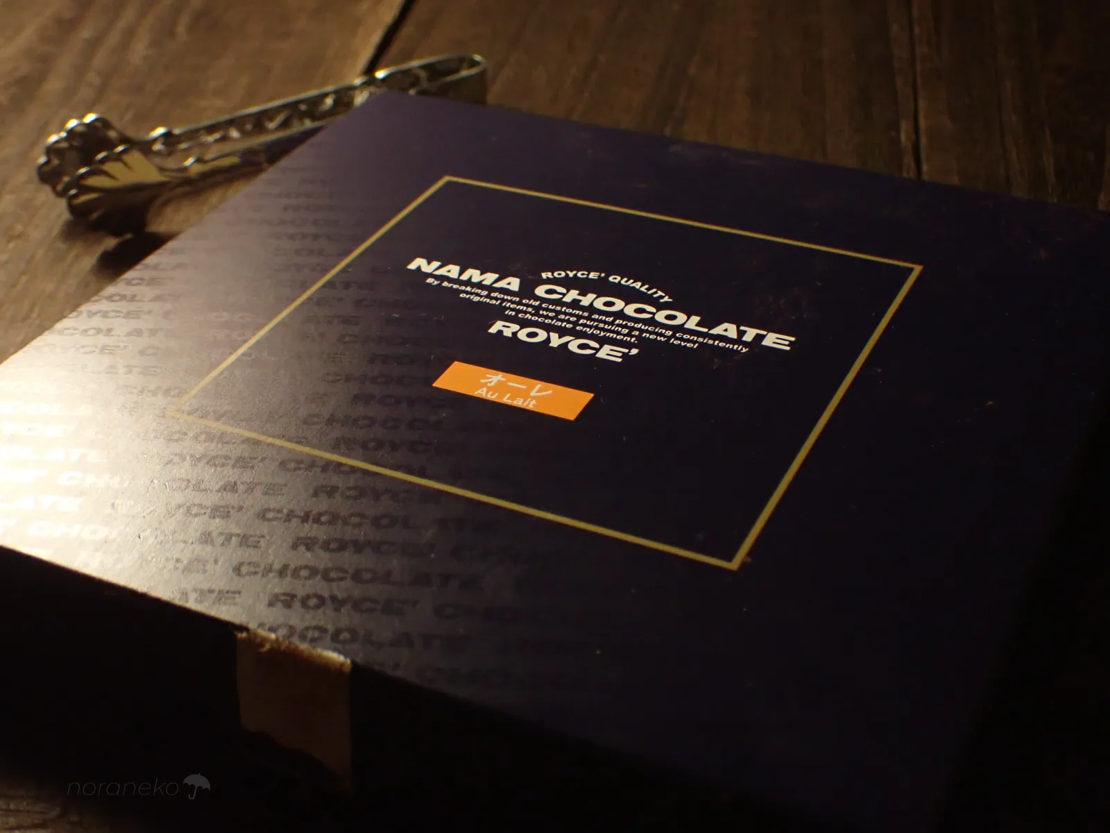 木目のテーブルに置かれた、ロイズ（ROYCE'）生チョコレート［オーレ］の濃紺のパッケージ。金色のロゴとフレーム、オレンジ色の小さなラベルが見える。左上にはシルバーのトング。