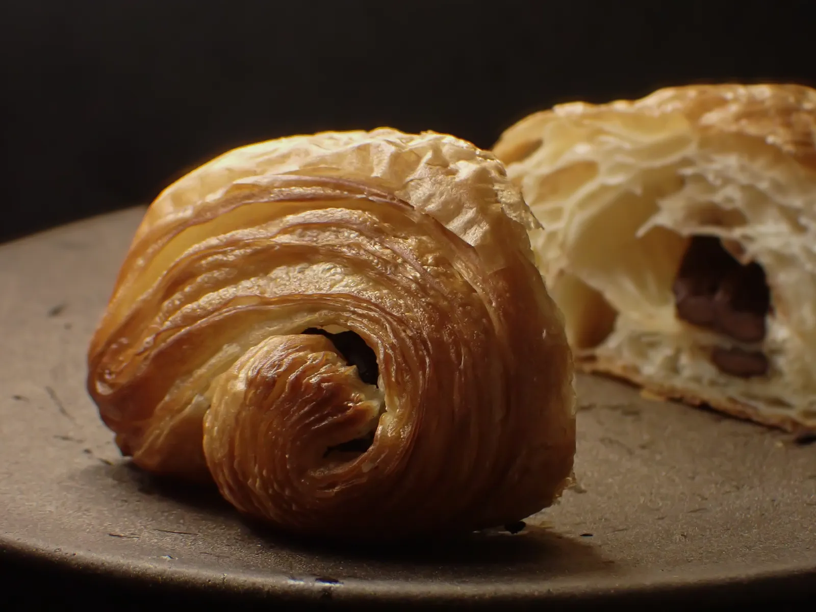 ベーカリーコネルのパンオショコラ。手前に焼き色の濃い渦巻く層、奥に淡い色の生地とチョコが見える断面を並べた写真。