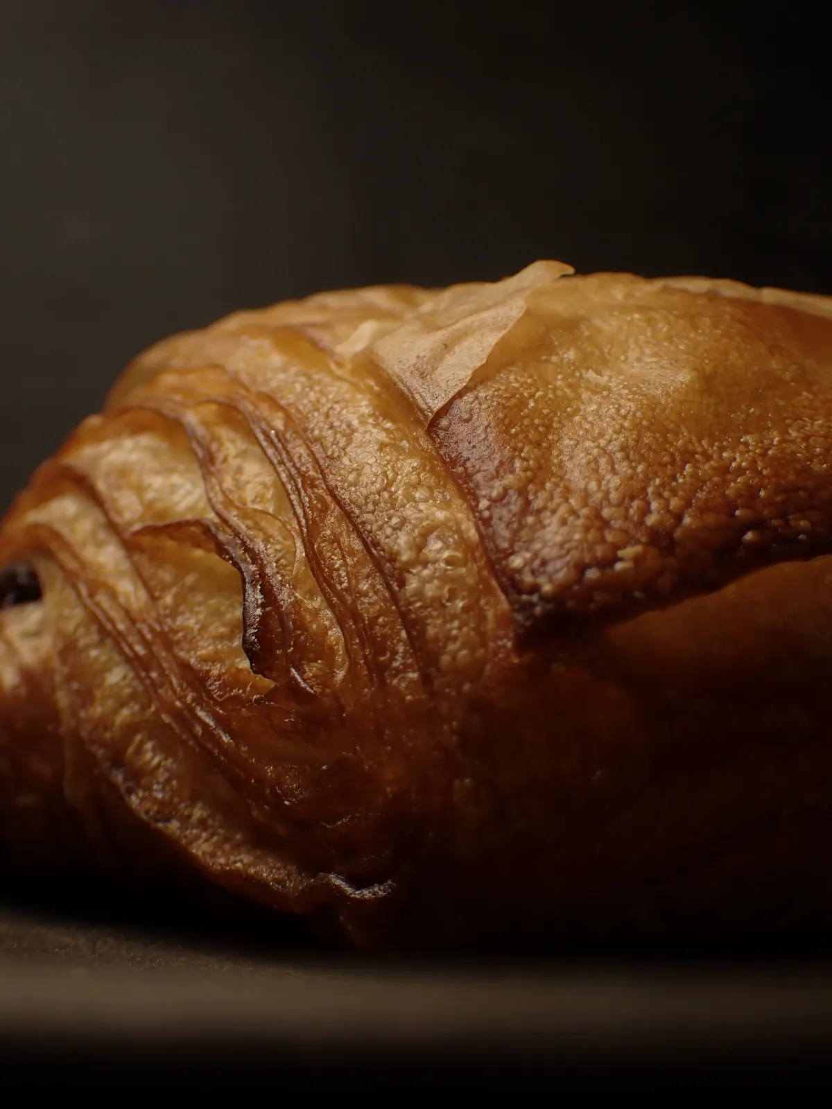 ベーカリーコネルのパンオショコラの表面を拡大。ところどころめくれた薄い皮と、焼き色の濃い層の縁、細かな凹凸が見える。